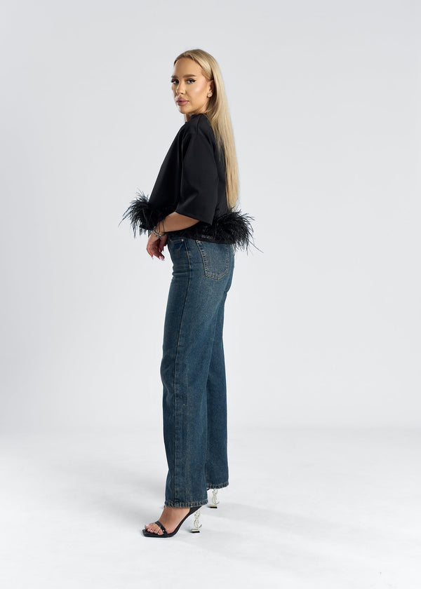 Femeie purtând un tricou mini oversized cu detalii din pene, model SIA Mini Feather Love, jeans și sandale.