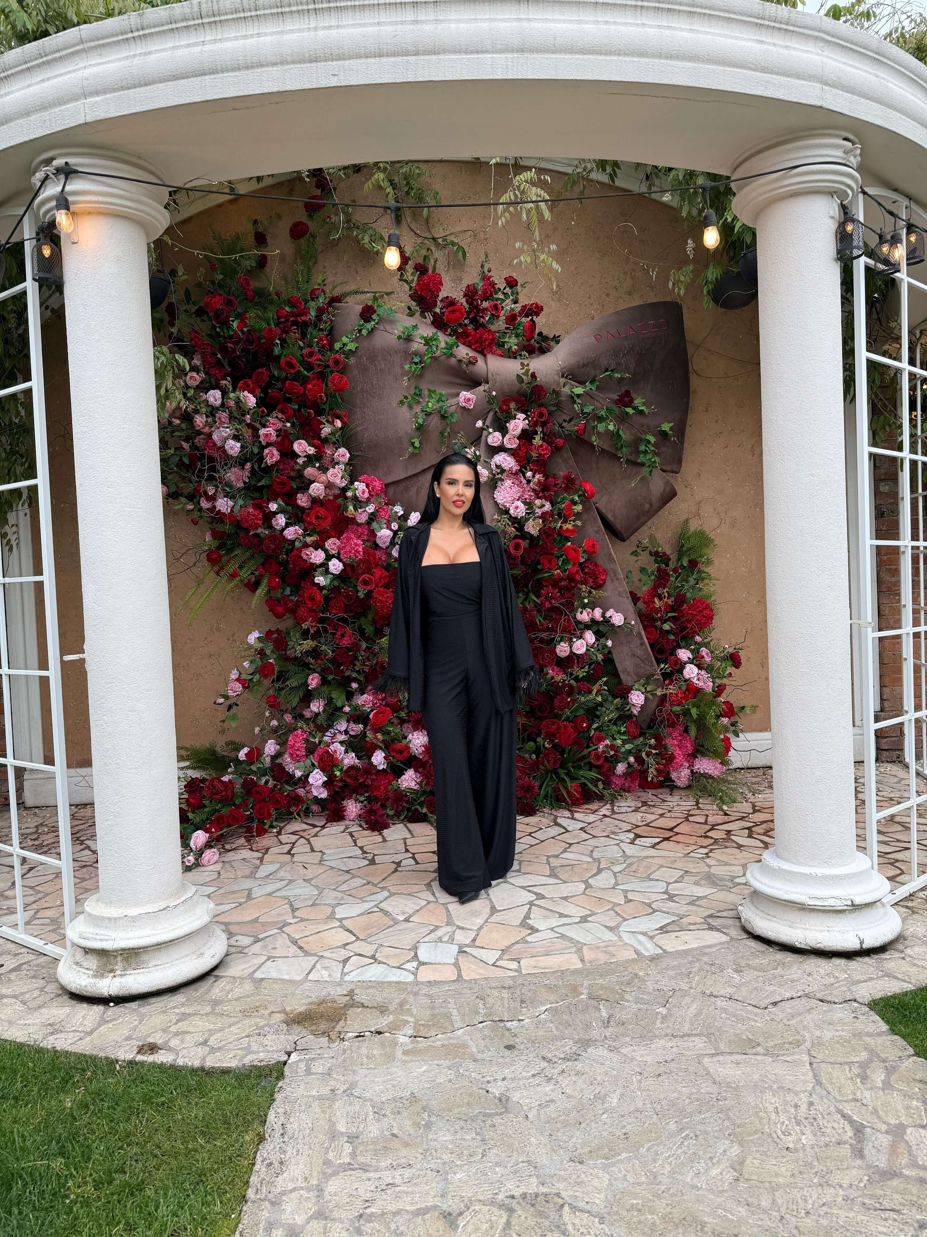 Femeie purtând Compleu Miray negru, format din bustieră, pantaloni evazați și mantou, pozată în fața unui decor floral elegant.